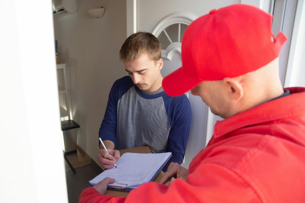 A delivery man in red uniform assists a young man signing for a package at the doorstep.